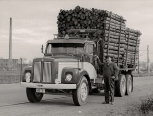 År 1961. Ett svart/vitt foto av grundaren Sven Jansson som står bredvid en blank ny Scania lastbil med flak. Flaket är lastat högt med timmer.